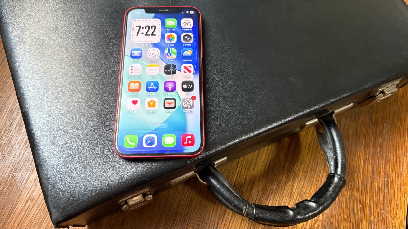 Red smartphone displaying app icons and time, resting on a closed black leather briefcase with a black handle on a wooden surface.
