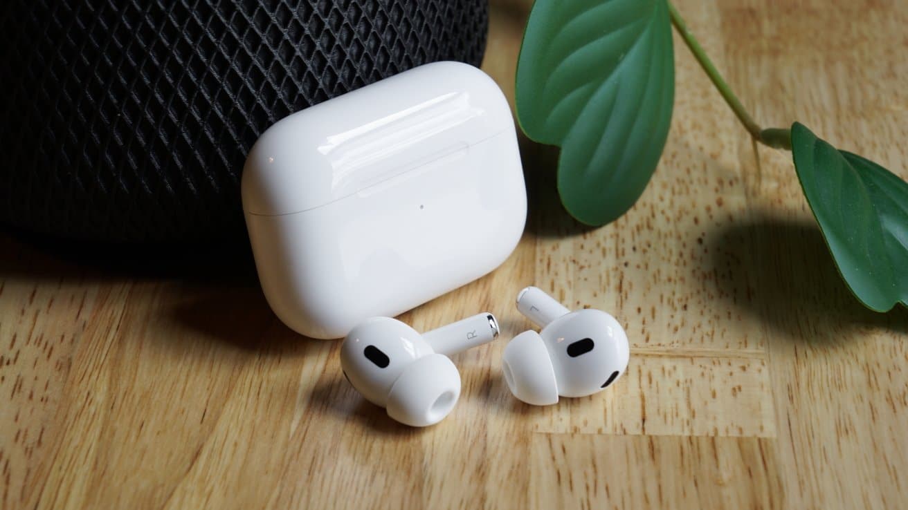 White wireless earbuds and charging case on a wooden surface with a dark textile and green leaves nearby.