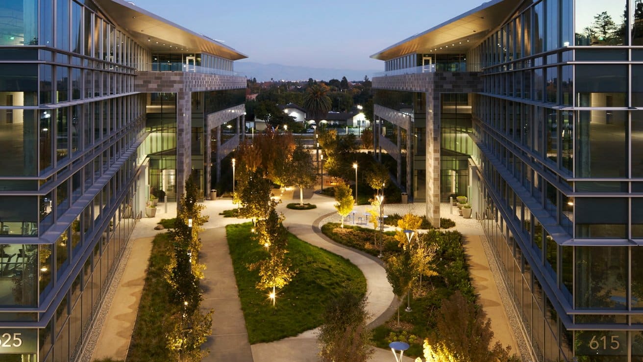 Modern office buildings with glass facades flank a landscaped courtyard featuring illuminated pathways, trees, and benches, creating an inviting atmosphere at dusk.