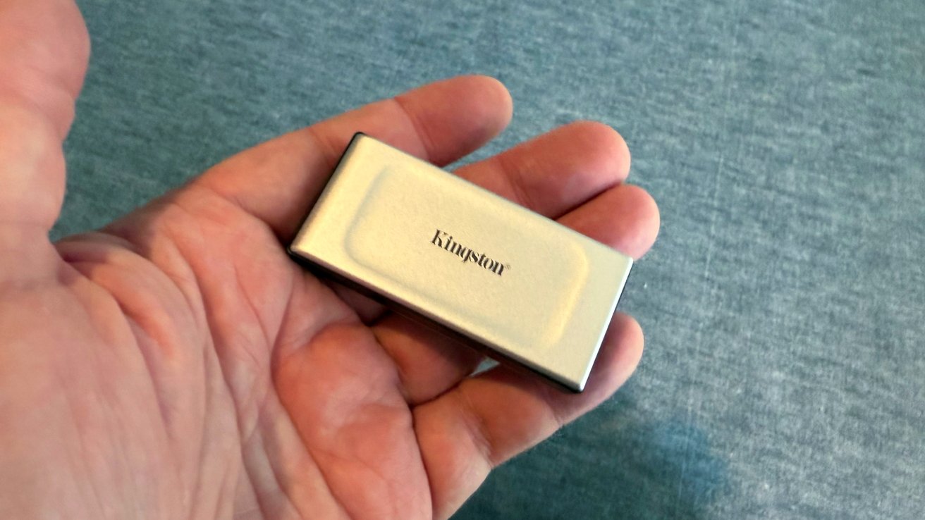A hand holds a small, rectangular metal device with 'Kingston' on its surface, set against a gray textured background.