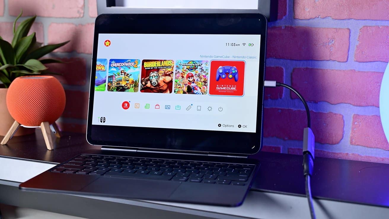 Tablet with a Nintendo game interface on a stand connected to a keyboard, on a table with a small orange speaker and potted plant, brick wall background.