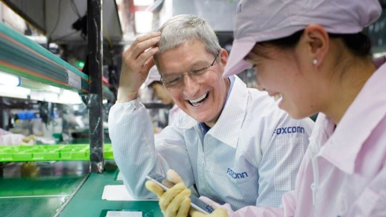 Two people in matching work attire, smiling and chatting at a workstation, with assembly line components around them.