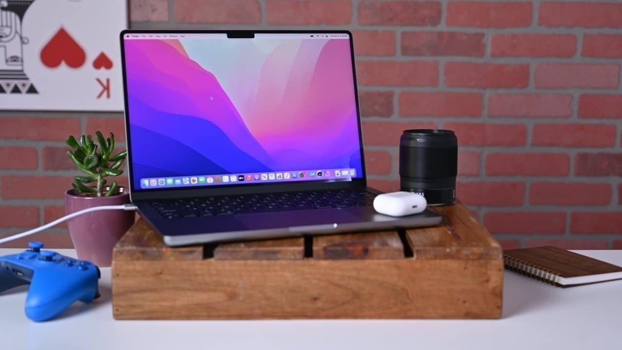 Laptop on wooden stand with colorful screen, small plant, blue gaming controller, camera lens, earphones, and notebook on white table against brick wall background.