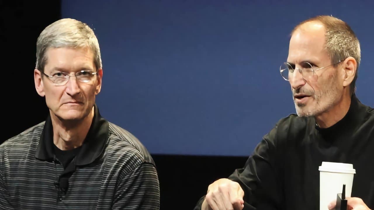 Two men with glasses sit together on stage; one wears a black shirt, the other a striped polo, holding a paper cup.