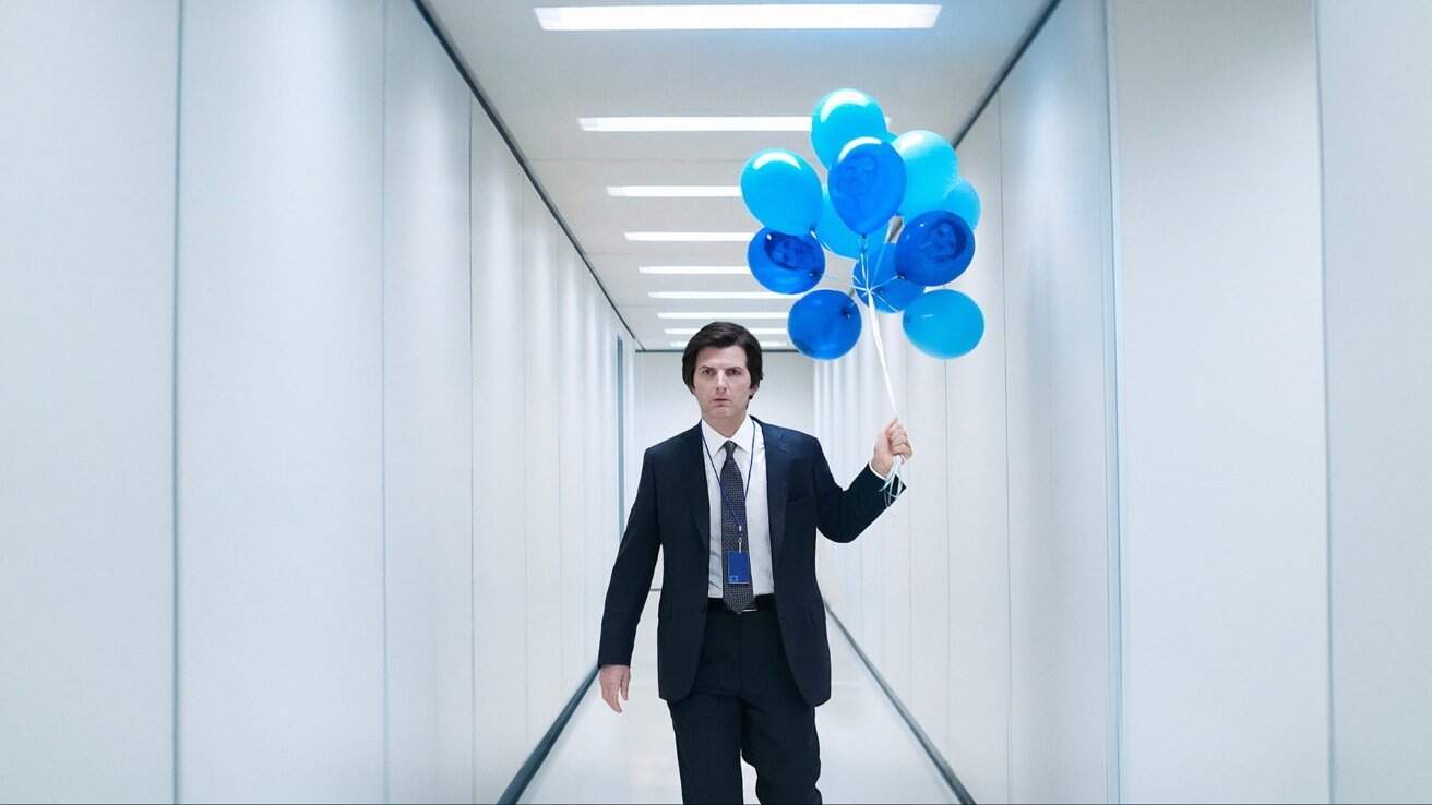 Man in suit holding blue balloons walks down a brightly lit, narrow corridor with white walls and ceiling lights.