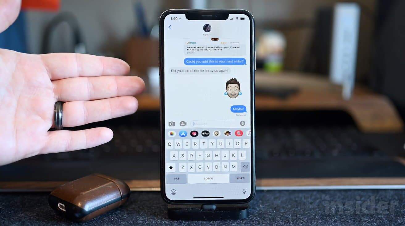 Hand gesturing near smartphone displaying a text conversation with emojis. Earbuds case nearby on a table with a blurred background.