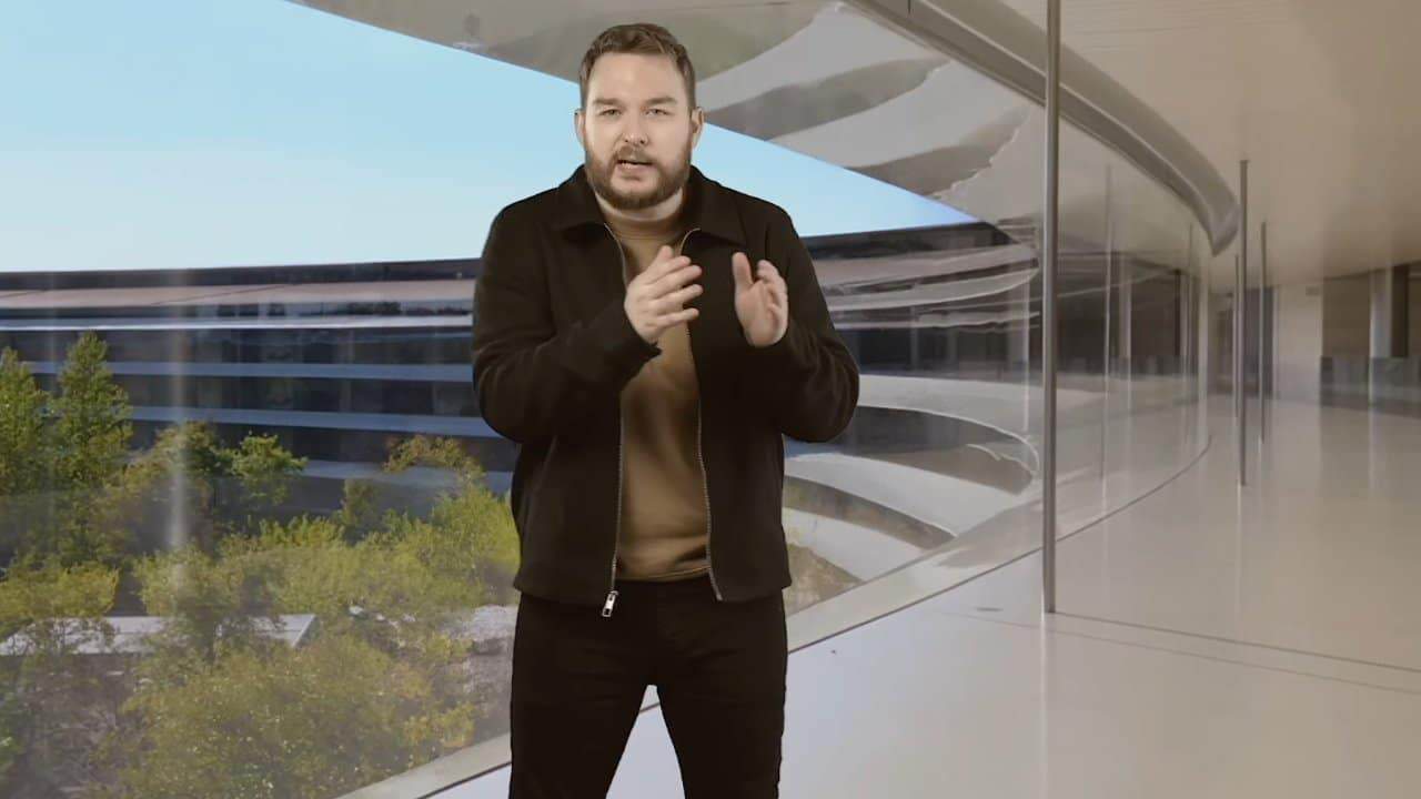 A man in a black jacket is gesturing while standing in a modern glass corridor with trees visible outside.