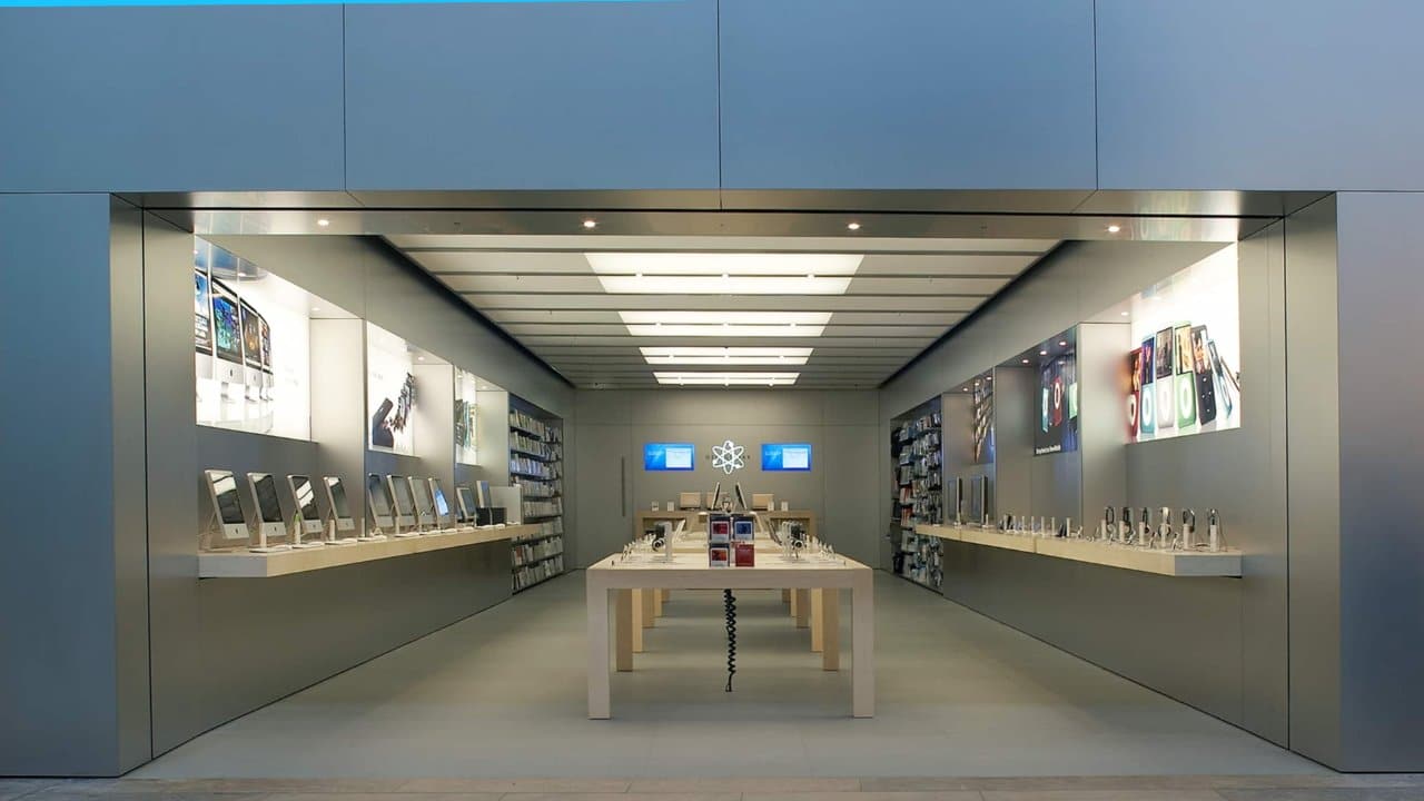 Modern electronics store interior with sleek gray walls, wooden tables displaying gadgets, accessories along the sides, and illuminated screens on the back wall.