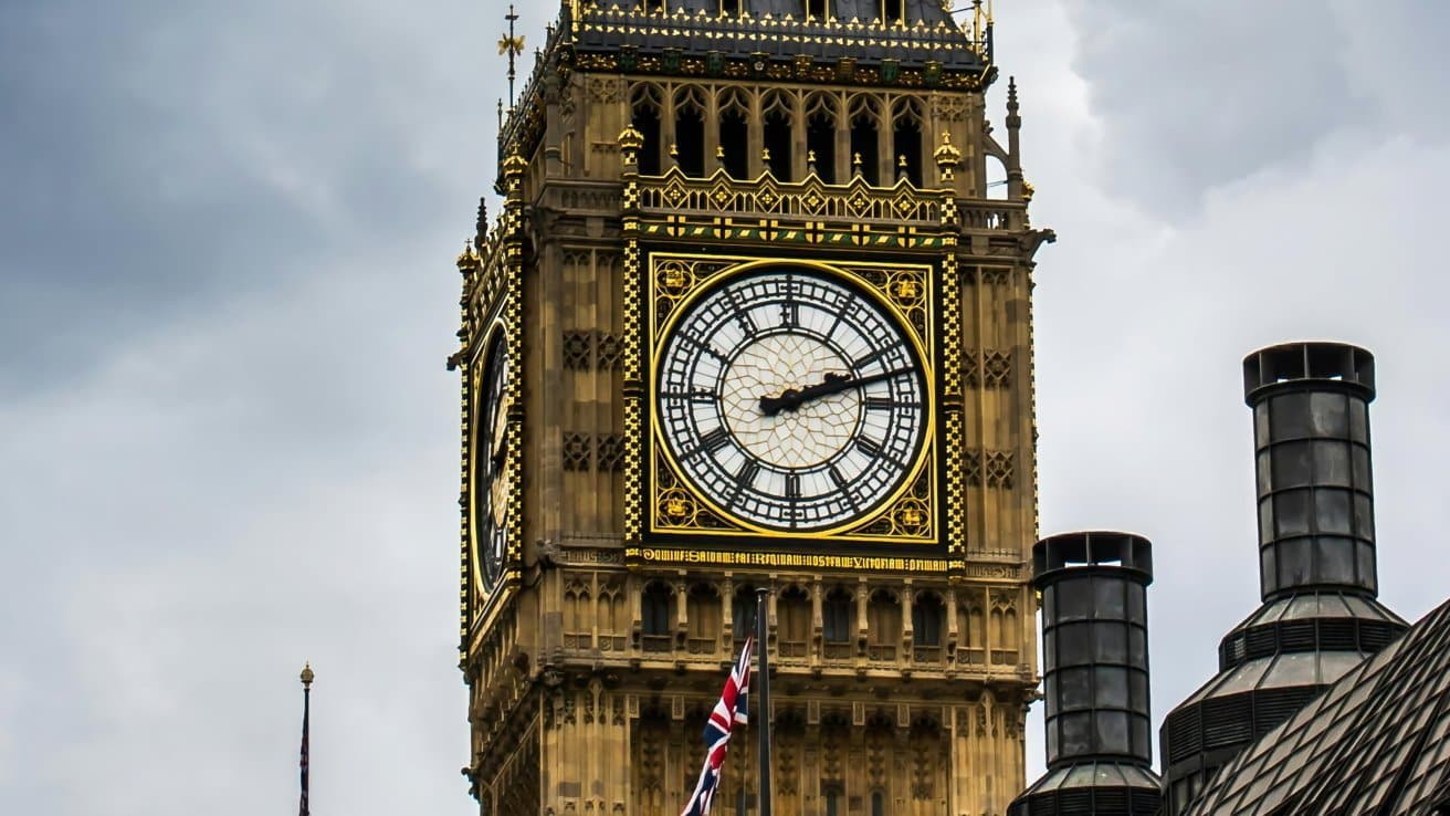Clock tower with ornate architecture, large white clock face, and golden accents; gray sky background. British flag visible below.