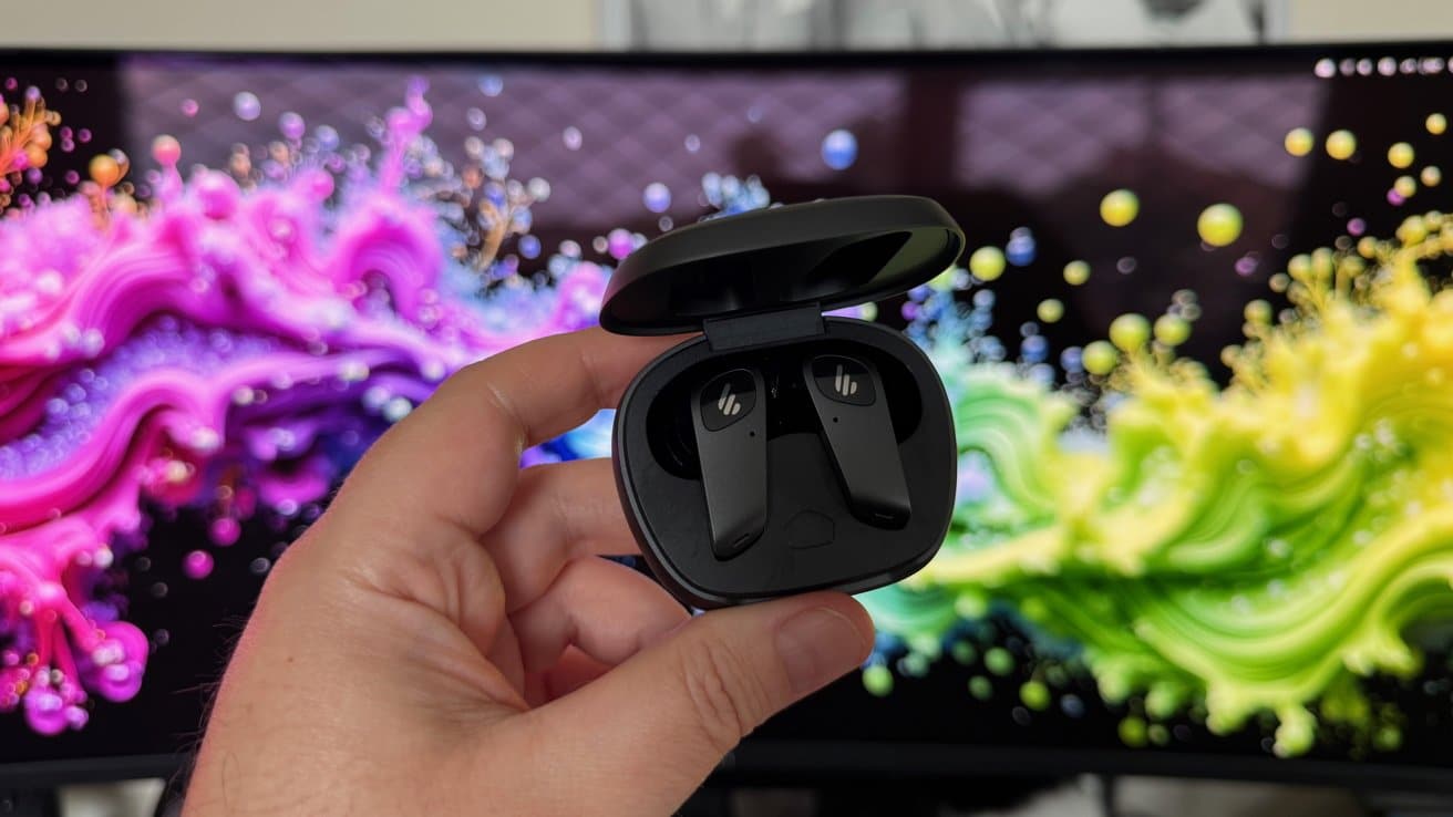Hand holding open case with black wireless earbuds, colorful swirling abstract background on monitor behind.