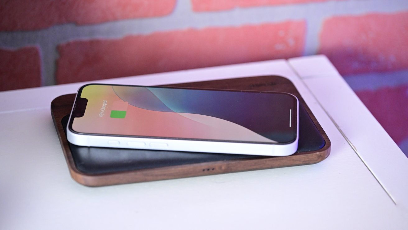 Smartphone charging wirelessly on a wooden charger atop a white surface, with a reddish brick-patterned background.