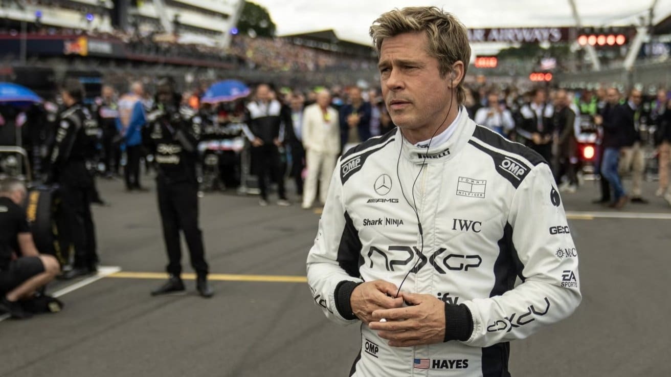 A man in a racing suit stands on a race track, surrounded by a crowd and pit crew members.