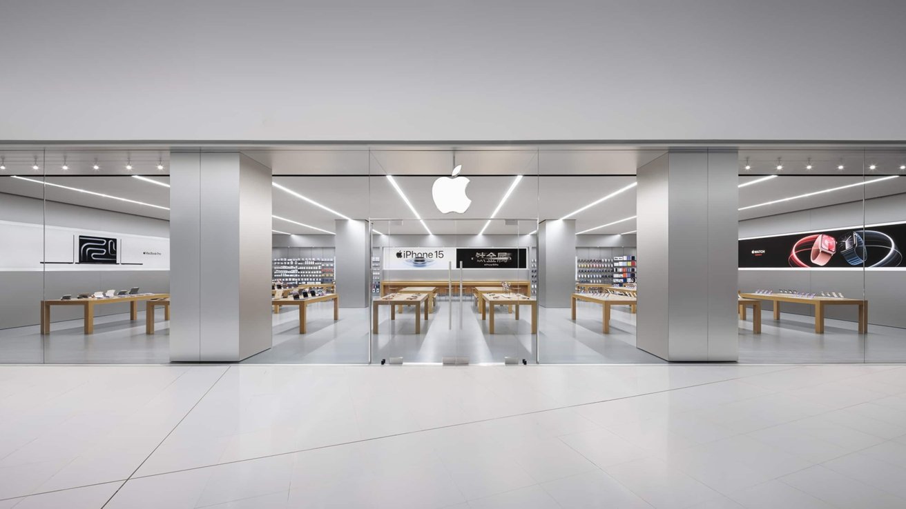Modern Apple store interior with wooden tables displaying products, sleek lighting, and illuminated Apple logo above glass doors.