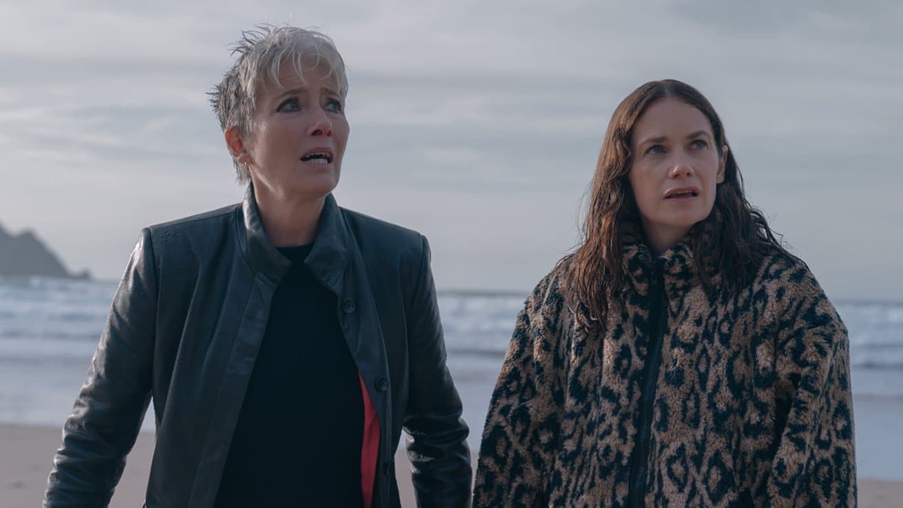 Two concerned women stand on a beach, one in a black jacket and the other in a leopard print coat, with waves and cloudy sky in the background.