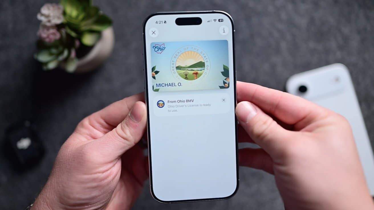 Hands holding a smartphone displaying a digital Ohio driver's license notification. Background shows a potted plant and a white device on a gray surface.