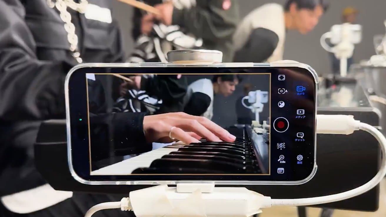 A smartphone on a stand captures a person playing a keyboard, with others in the blurred background.