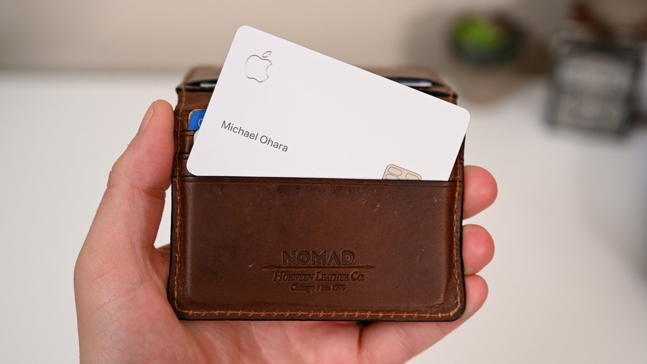 Hand holding a brown leather wallet with a white card partially visible, featuring an embossed Apple logo and text.