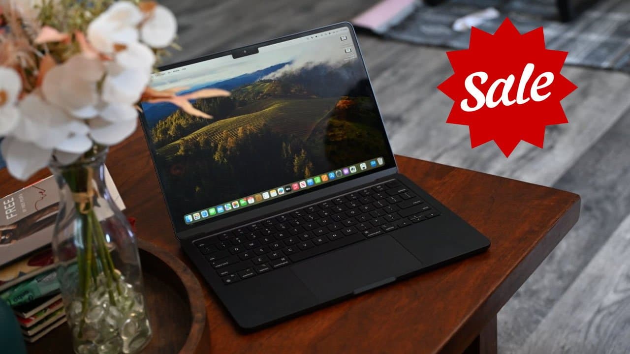 MacBook Air in Midnight finish on wooden table with a scenic screen, surrounded by books and a vase of flowers; red sale badge on the right.