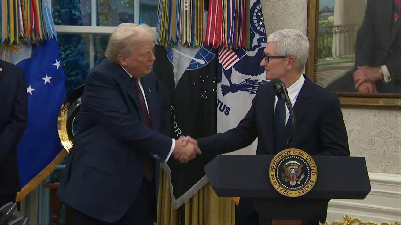 Two men in suits shake hands in an official setting, with flags and a painting in the background.