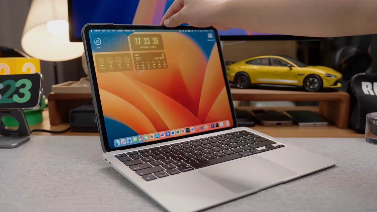 Laptop with colorful screen on a desk, hand touching top edge, yellow toy car in background, various items around.