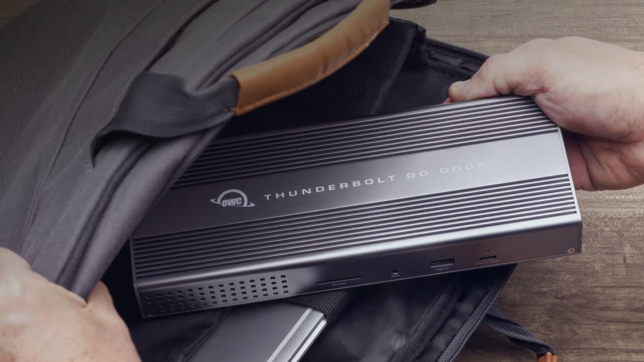 A silver Thunderbolt Go Dock is being placed into a gray backpack with leather accents by a person on a wooden surface.
