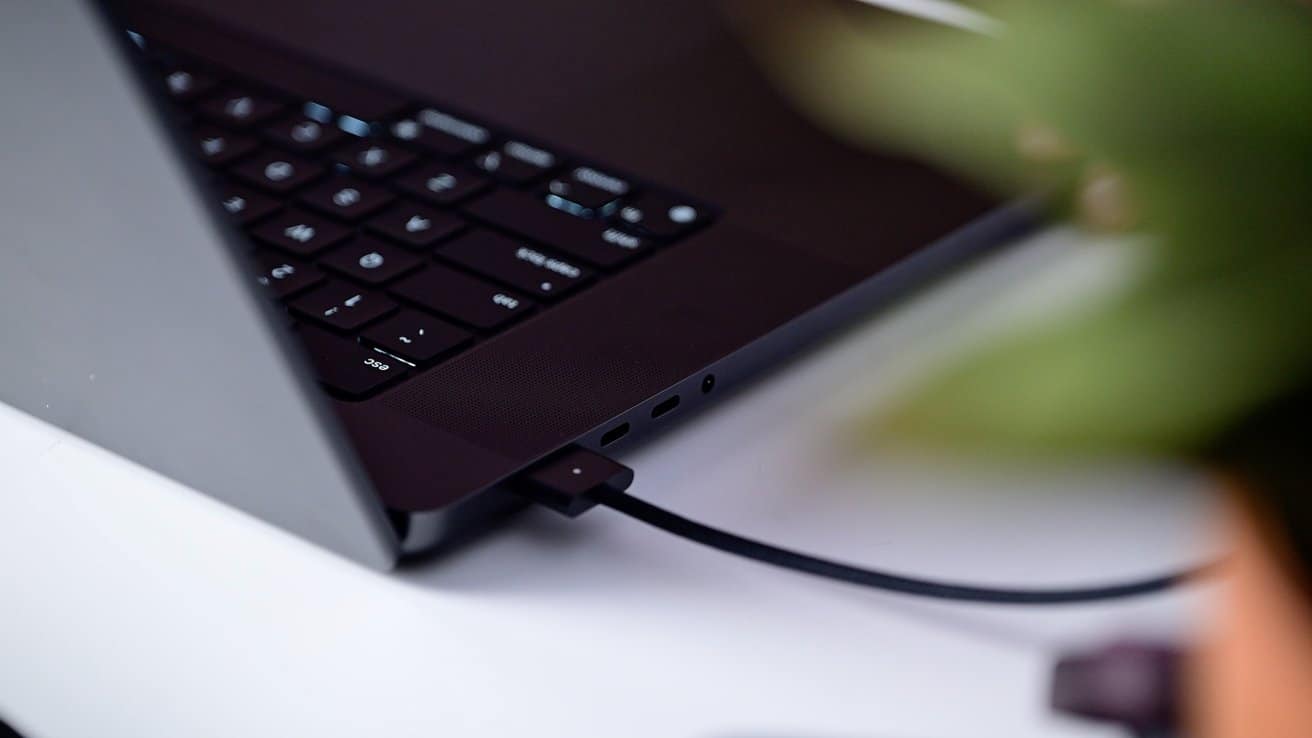 A close-up view of a laptop with a USB cable connected, positioned on a white surface. The background is blurred with a hint of a plant.