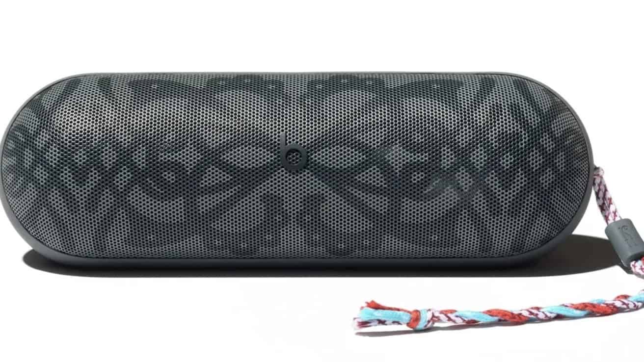 Black portable speaker with patterned front and rounded edges, featuring a woven multicolored strap attached on the right side.