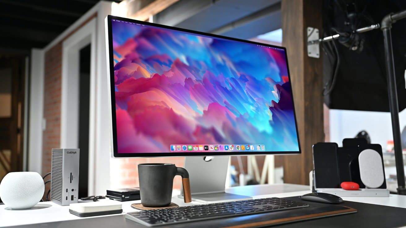 A desktop setup featuring a large monitor with vibrant abstract art, keyboard, mouse, headphones, mug, and tech devices on a white desk in a modern workspace.