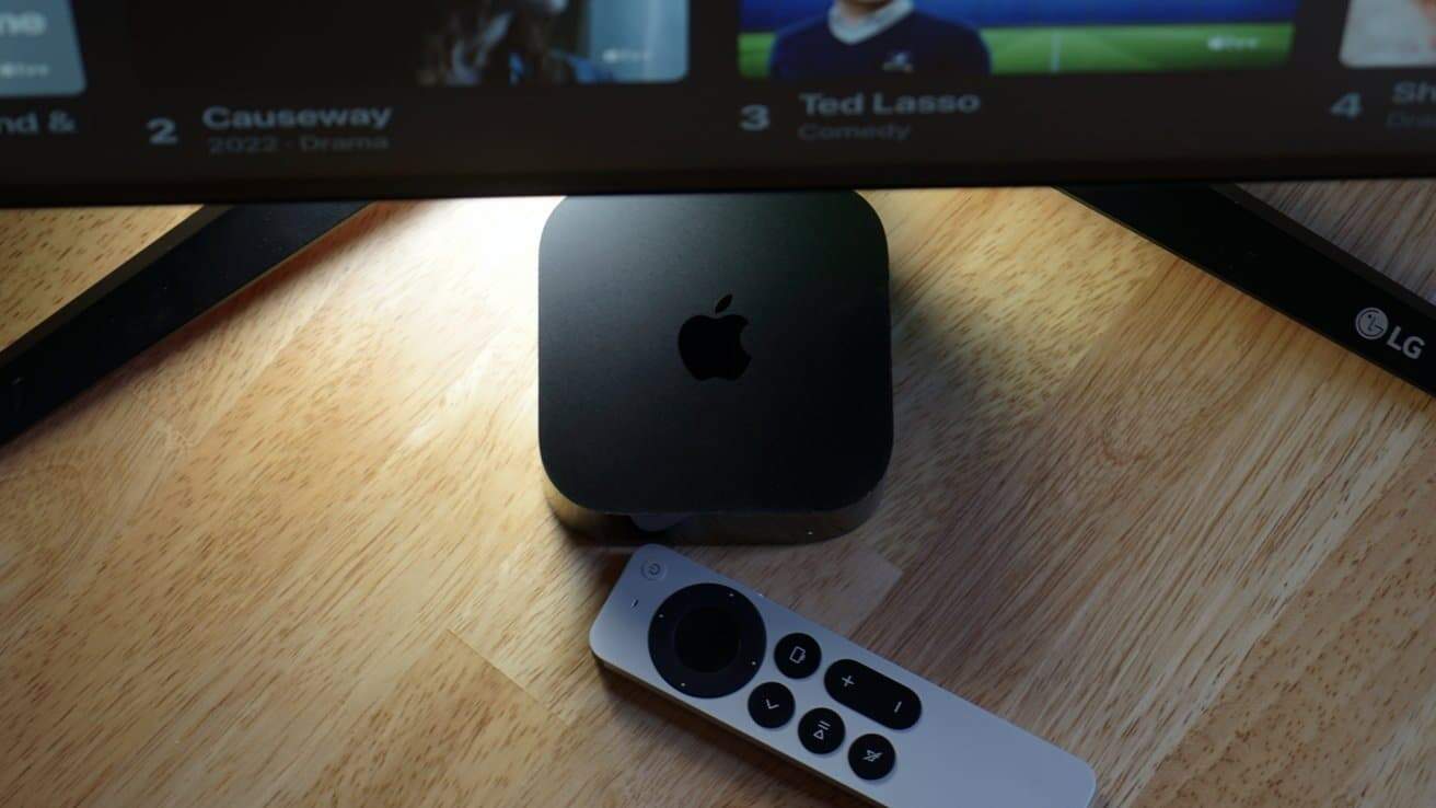 A black Apple TV box, remote control, and television screen on a wooden table; partial screen displays show titles and an LG logo.