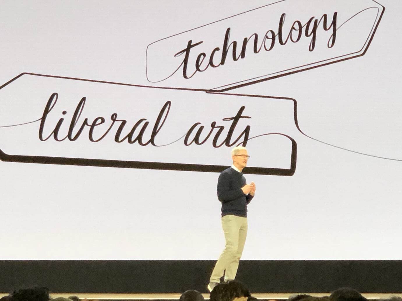 A person stands on a stage in front of a large screen displaying 'technology' and 'liberal arts' in handwritten style.