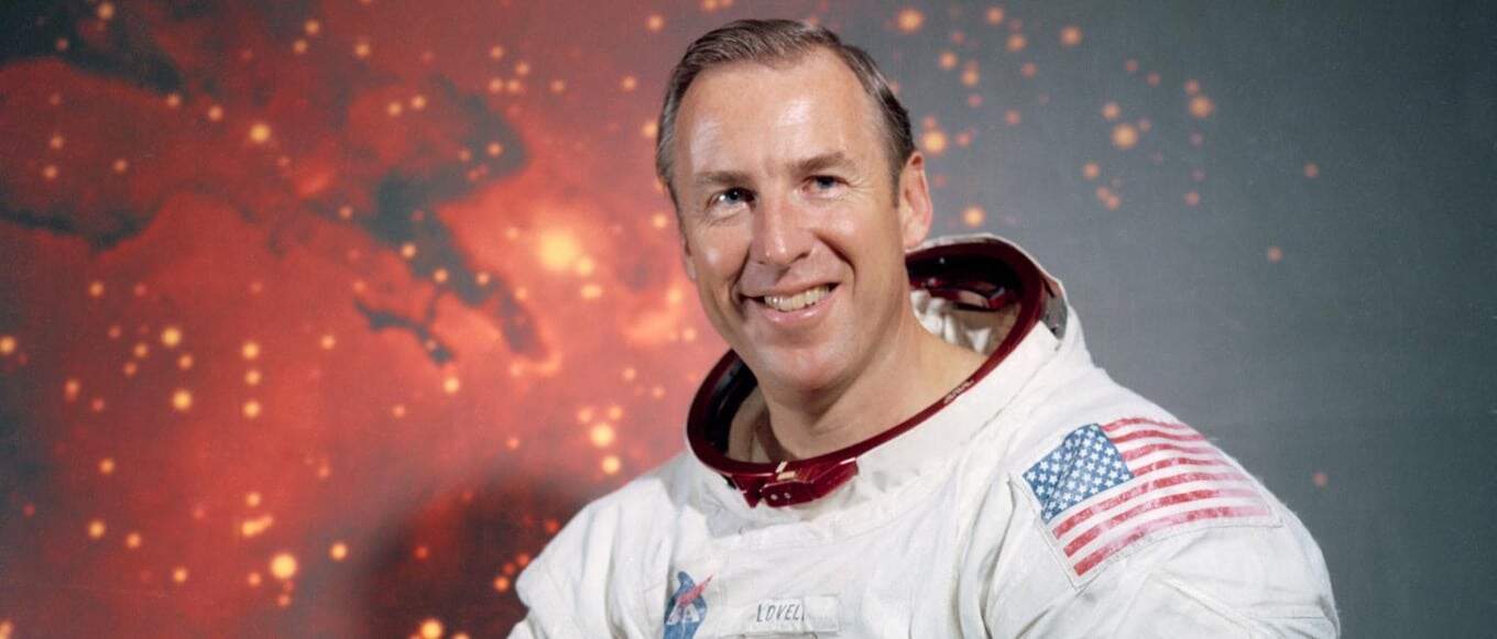 A man in a white astronaut suit with an American flag patch smiles against a cosmic background of red and orange nebulae and stars.