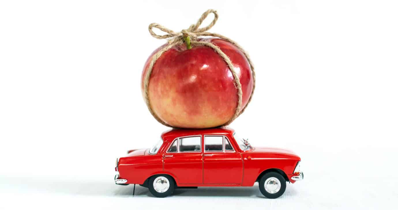 A red toy car balances a large red apple tied with twine on its roof against a white background.