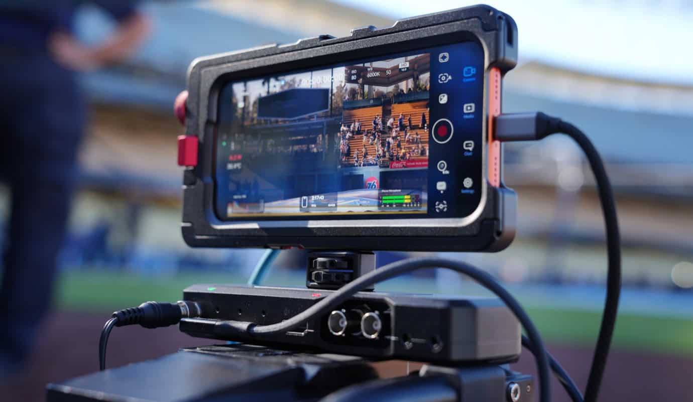 A smartphone mounted on a camera rig displays a live sports event. The screen shows a stadium with spectators and a field. The rig includes cables and a battery pack for power.