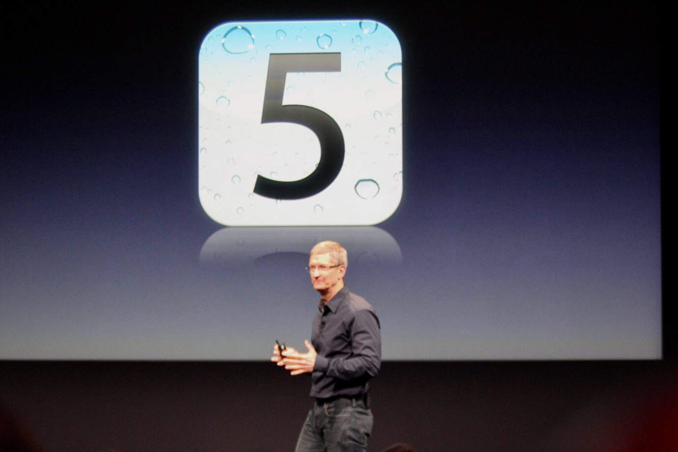 A man in a dark shirt stands on stage in front of a large screen displaying an 'iCloud' icon with the number '5.'