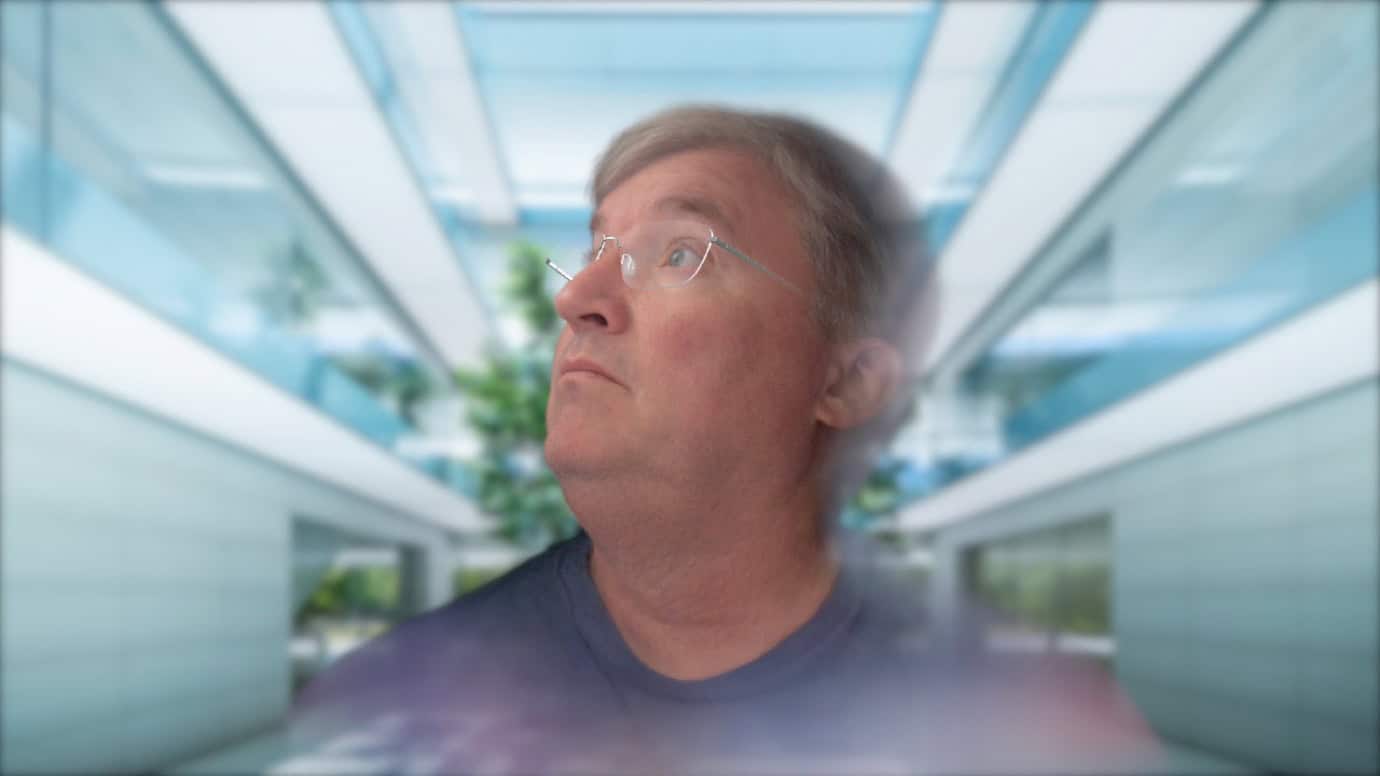 A man with glasses looking up in a modern, glass-roofed corridor with plants and bright lighting.
