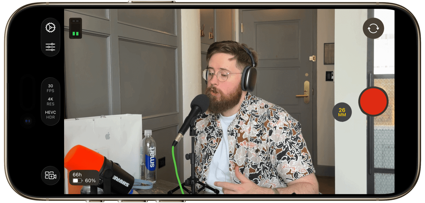 A man with a beard and glasses wearing headphones sits at a desk with a microphone and a water bottle. A smartphone screen shows a video recording interface with settings like '30 FPS,' '4K,' and a red record button. '26 MM' is displayed.