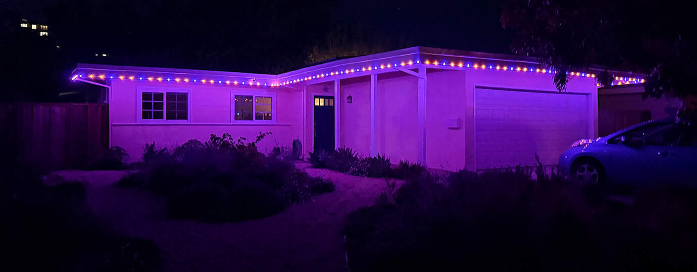 A house illuminated with purple string lights at night. A car is parked in the driveway, and shrubs line the front yard.