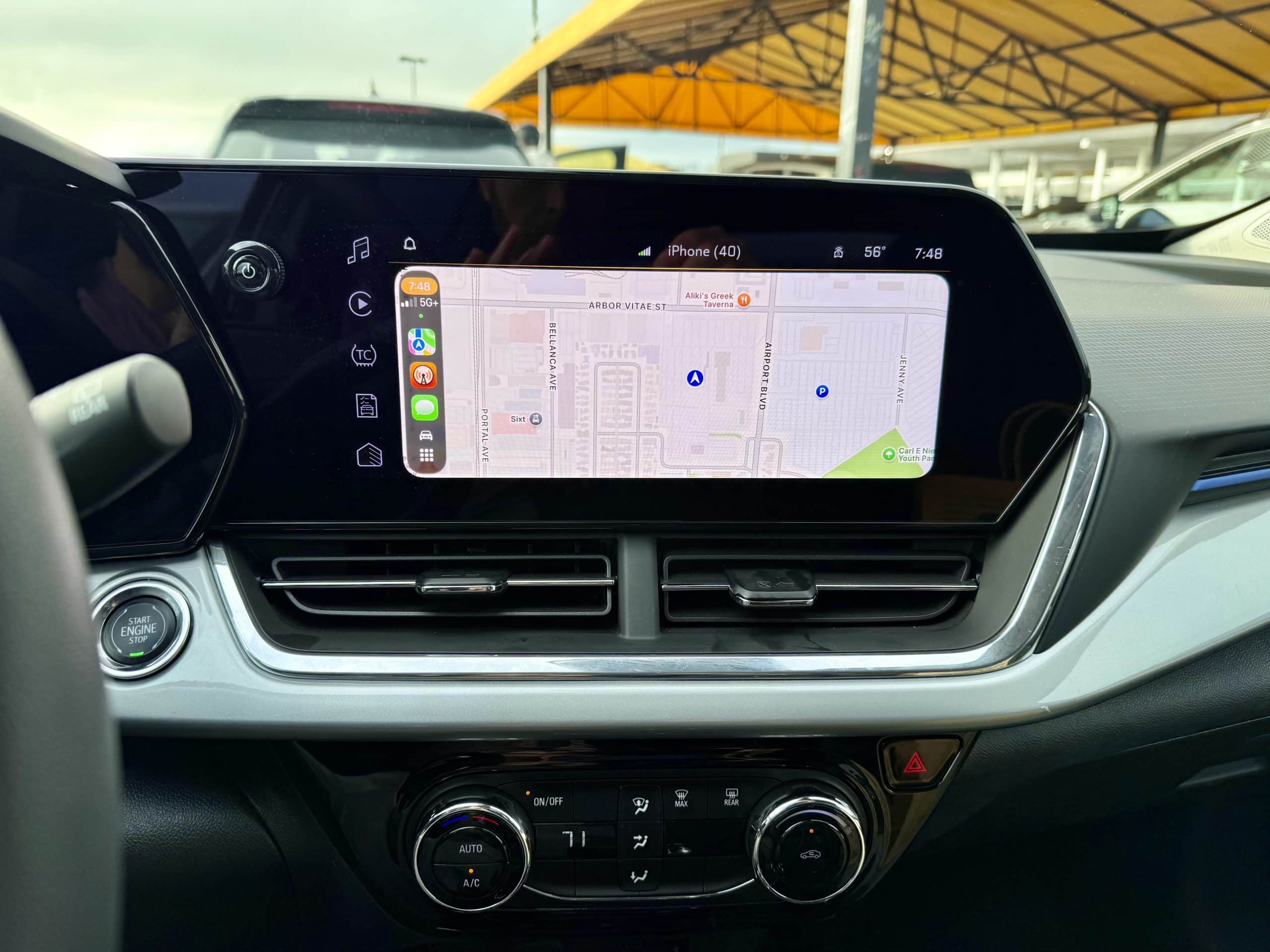 A photo of the center screen and air conditioning stack in a Chevy Trax. The screen displays the CarPlay maps interface. The other side of the windshield is the busy rental return center and awnings.