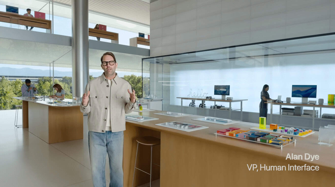 A man in a beige jacket stands in a modern, bright store with wooden tables displaying tech products. Glass walls reveal a scenic view. Text: 'Alan Dye, VP, Human Interface.'