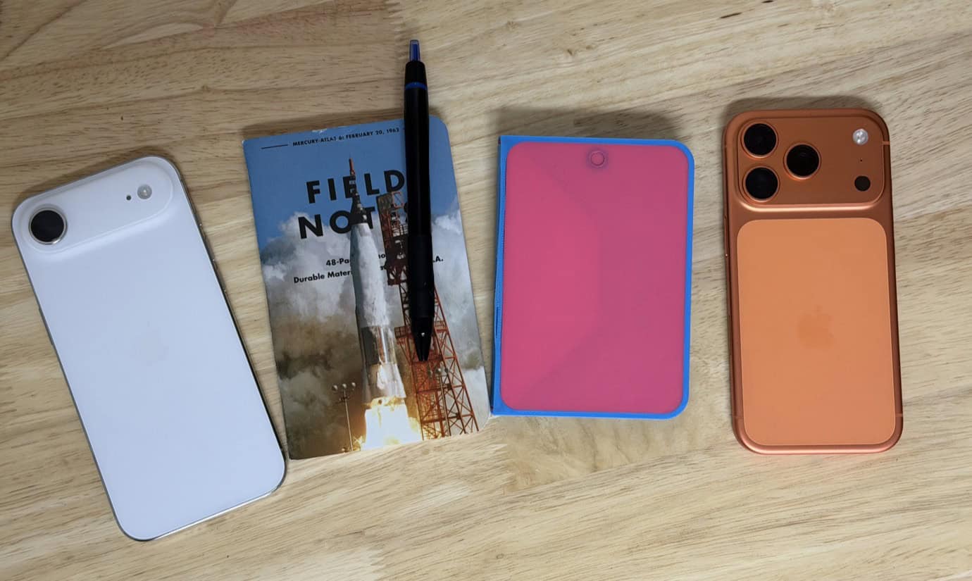 A white phone, a blue-bordered pink case, and an orange phone case with three camera lenses on a wooden surface. Next to them is a book titled 'Field Notes' with a rocket launch image and a black pen.
