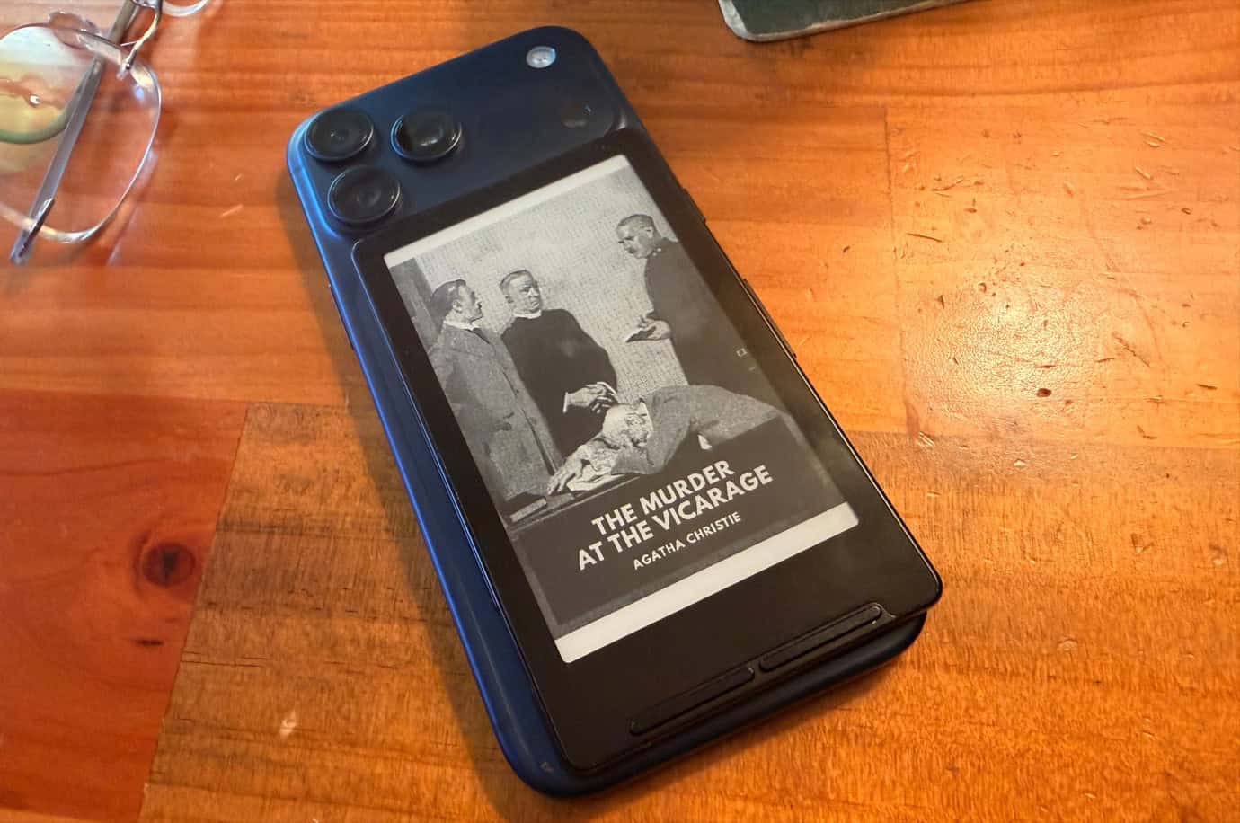 An iPhone with an Xteink X4 sitting on its back, displaying a black-and-white image of 'The Murder at the Vicarage' by Agatha Christie.