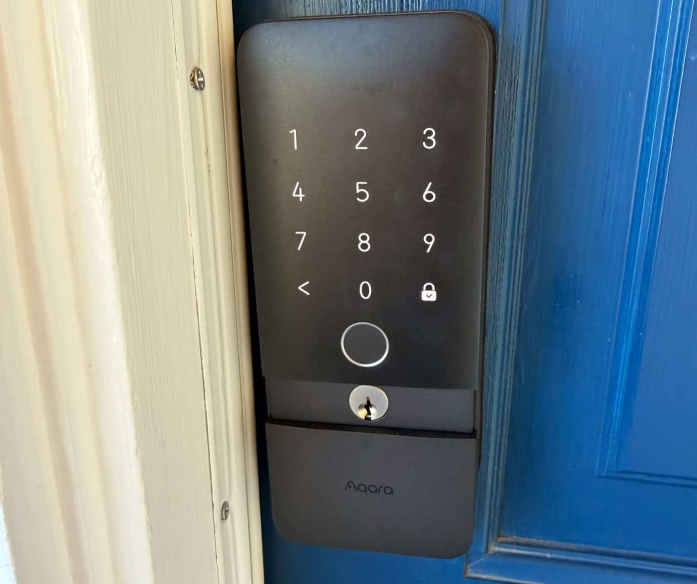 Black smart lock with white keypad on blue door. Keypad displays numbers 1-9, 0, and lock/unlock icons. 'Aqara' logo below keypad.