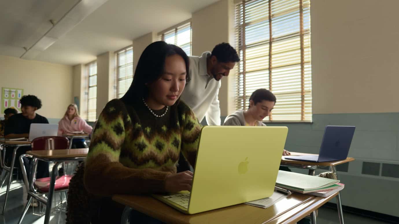 A classroom with students using laptops. A woman in a green sweater works on a yellow laptop. A teacher leans over a student's desk. Other students are seated at desks with laptops.