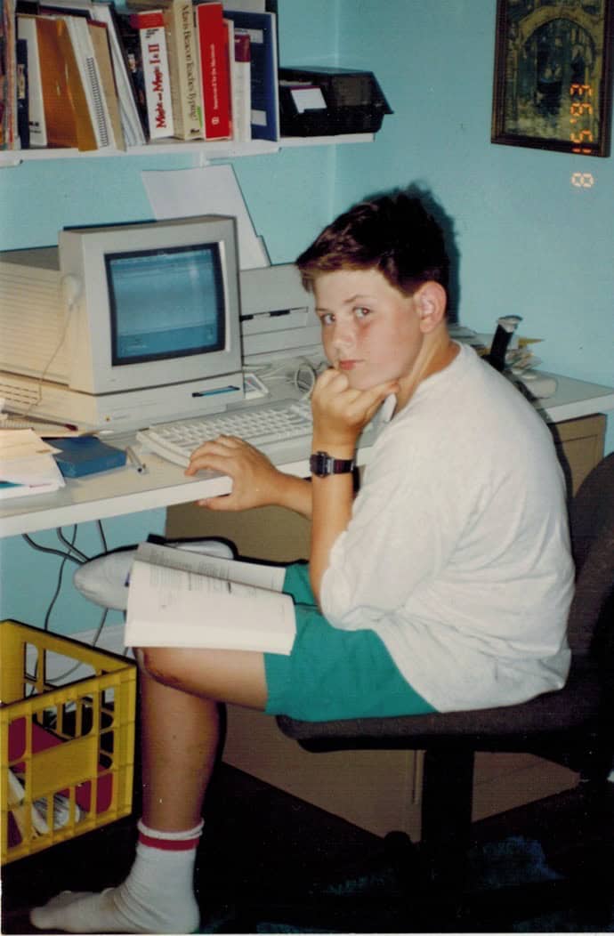 Thirteen-year-old Dan sitting at a Macintosh LC with a book open on his lap.