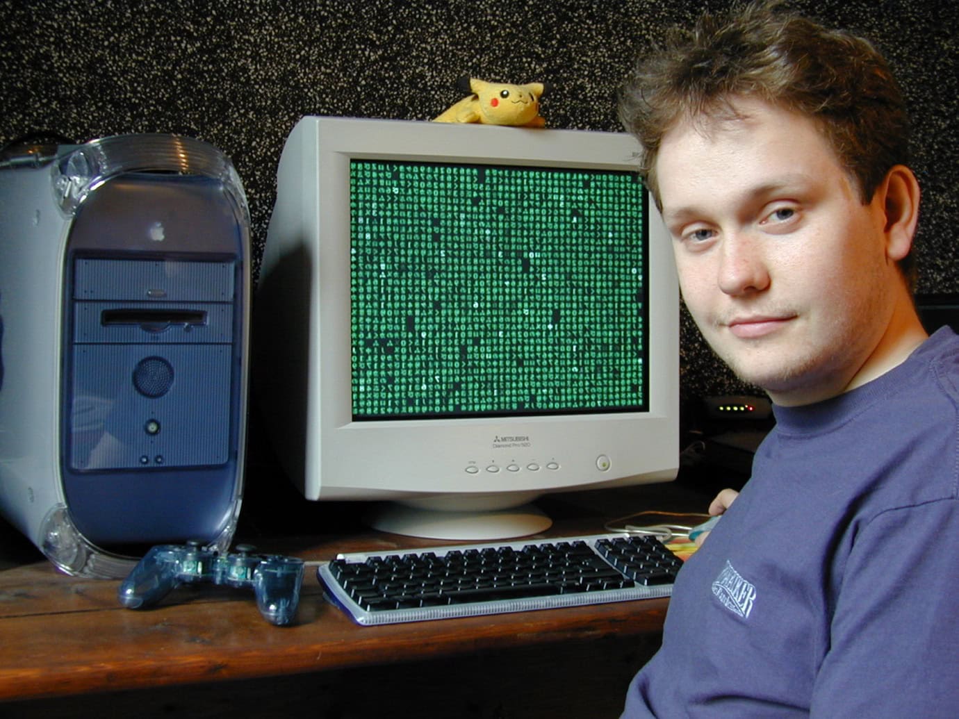 A man poses next to a vintage computer with a green Matrix-style screen, a PlayStation controller, and a Pikachu figurine on top. The setup is on a wooden desk against a speckled wall.