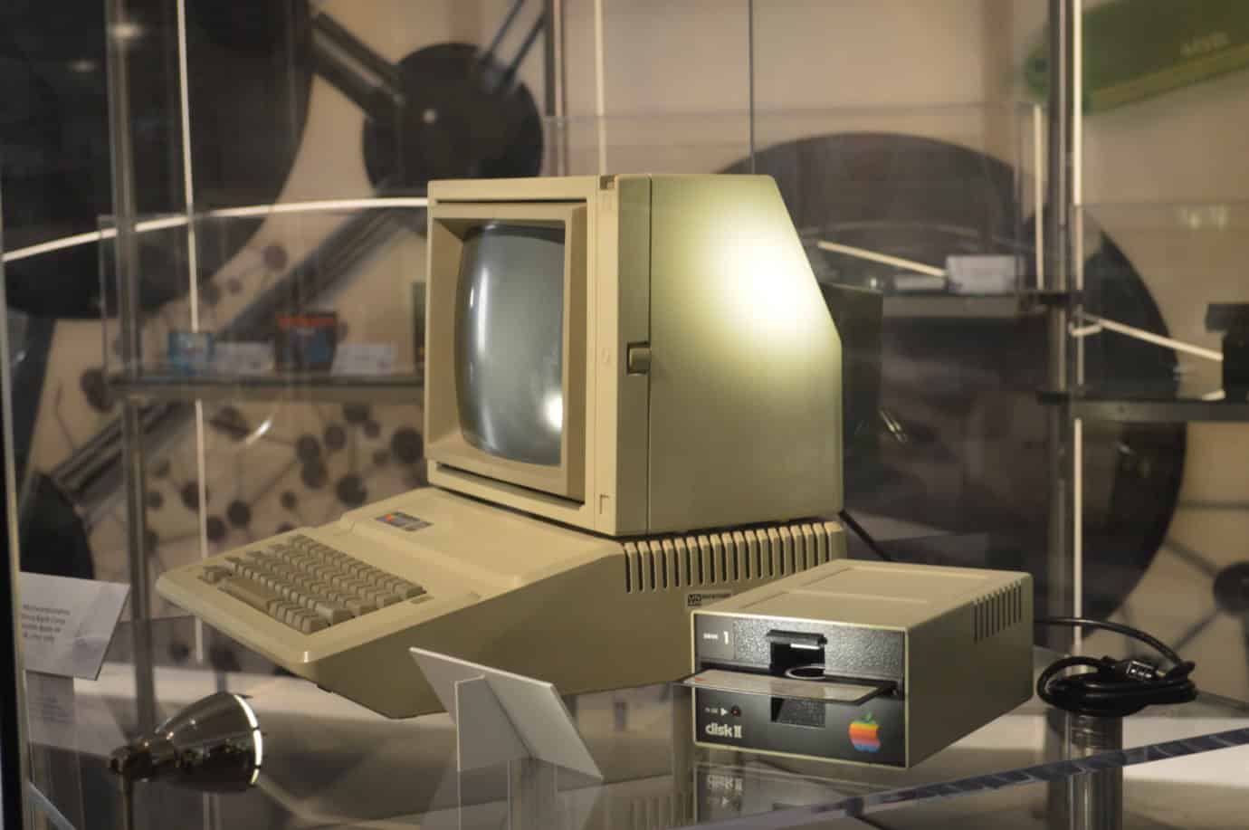 Vintage Apple II computer with a beige monitor, keyboard, and floppy disk drive in a glass display case.